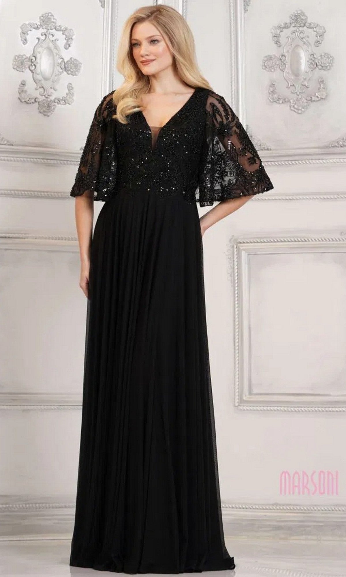 A woman wearing the black Long Formal Dress: Marsoni MV1302, featuring a floor-length silhouette, beaded details, deep V-neckline, lace sleeves, and sequined bodice stands against an ornate white wall.